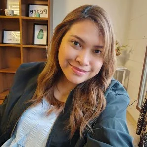 Meet the Team: A person with long hair smiles beside a bookshelf indoors.