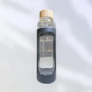 A sleek promotional glass water bottle with a wooden cap is enclosed in a black silicone sleeve, featuring an oval cutout. The soft, blurry background adds an elegant touch to its sophisticated design.