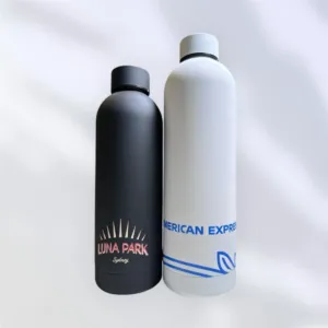 Two promotional bottles sit side by side. The black bottle on the left features Luna Park Sydney in bold white and red text, while the white bottle on the right sports the distinctive blue logo and text of American Express. Both are equipped with screw-on caps.