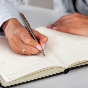 A person is writing in an open planner with a pen, focusing on a few blank pages of their branded corporate diary.
