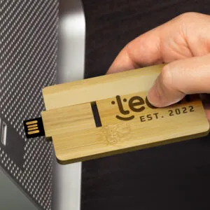 A hand holds a custom USB drive, elegantly crafted from wood and engraved with EST. 2022, as its plugged into a computer. Promote in style with these sophisticated devices that blend form and function seamlessly.