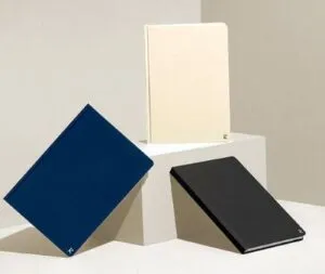Three notebooks in blue, beige, and black are displayed on a minimalist platform against a neutral background, perfect products for conference promotions.