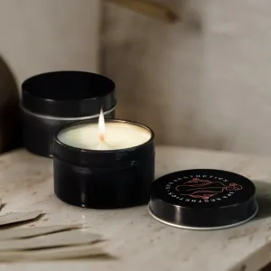 Two black tin candles sit elegantly on a table; one is open and lit, casting a warm glow, while the other remains closed. Part of Sheas Favourite autumn merch, these candles bring cozy charm to any setting.
