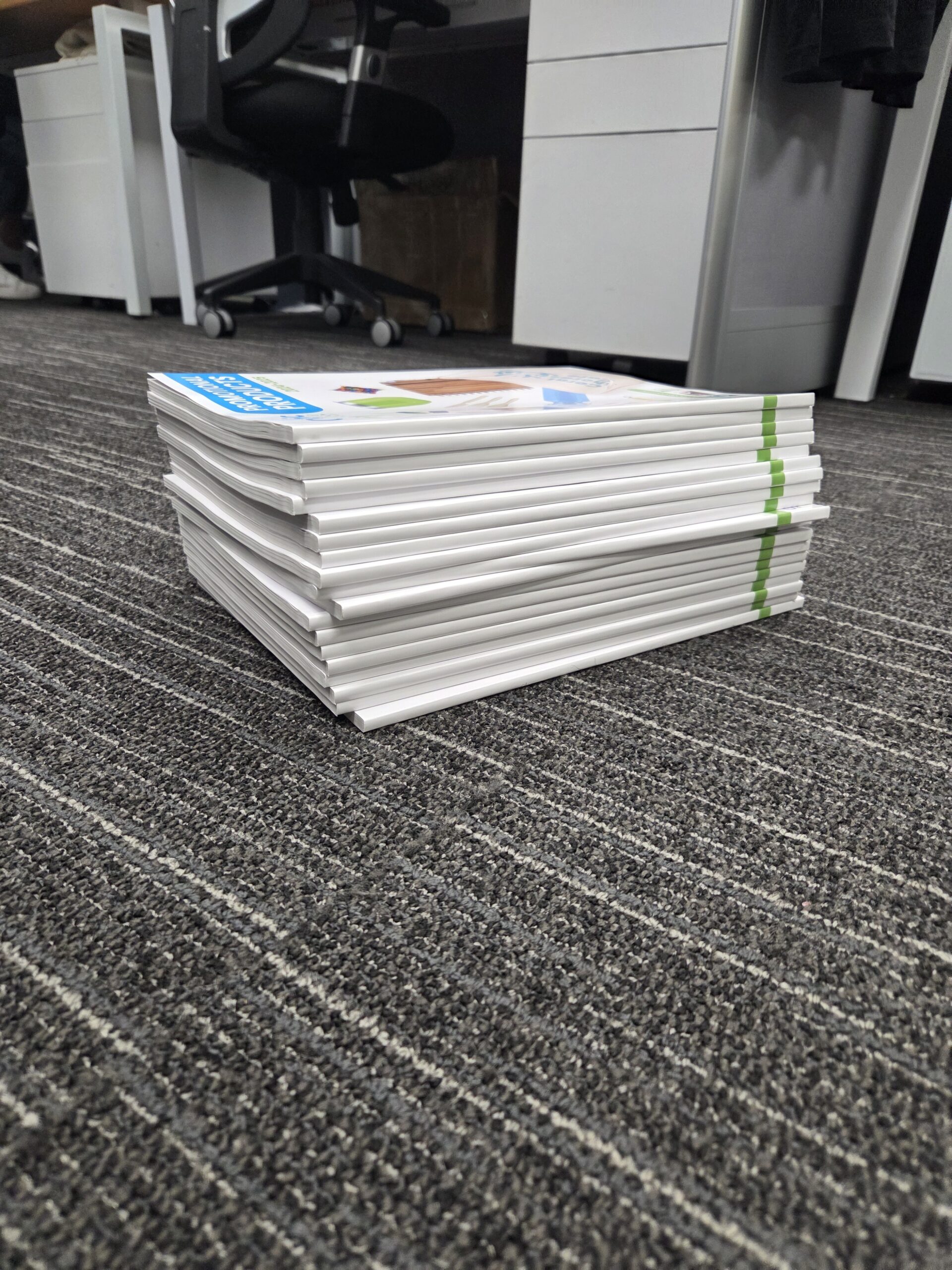 A stack of books or magazines rests near desks, beside conference bags on the gray carpeted floor.