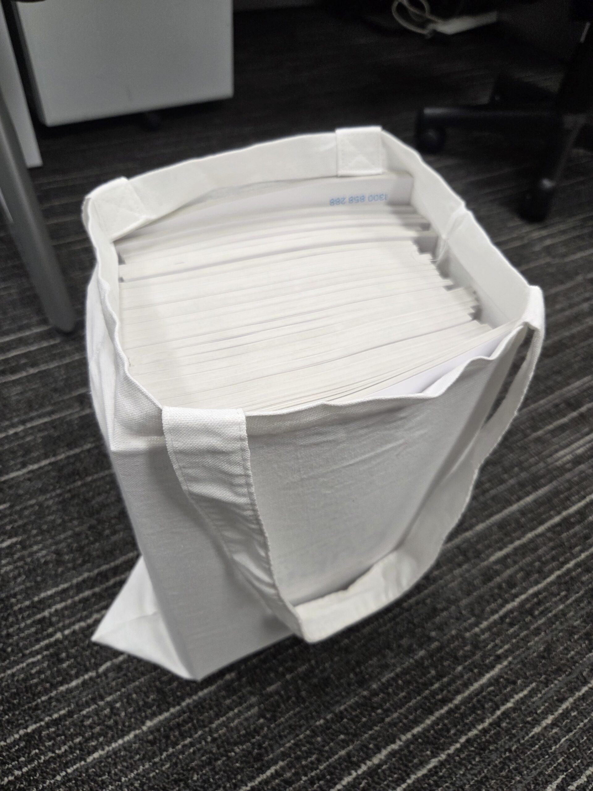A white conference bag filled with a large stack of white envelopes on an office floor.