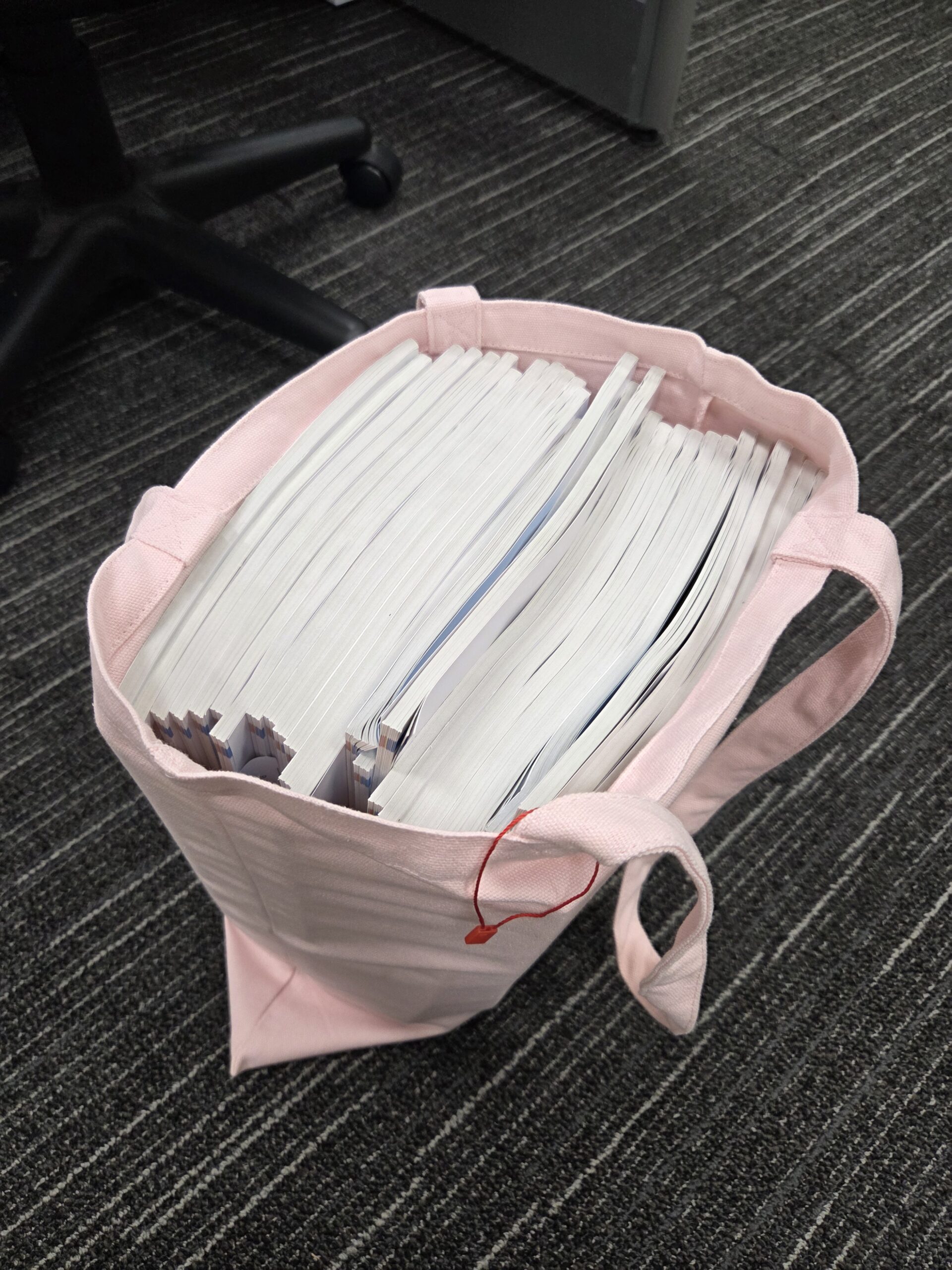 A pink conference tote bag brimming with white documents rests on a carpeted floor.