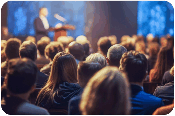Audience seated in a dimly lit room, listening to a conference speaker at a podium on stage.