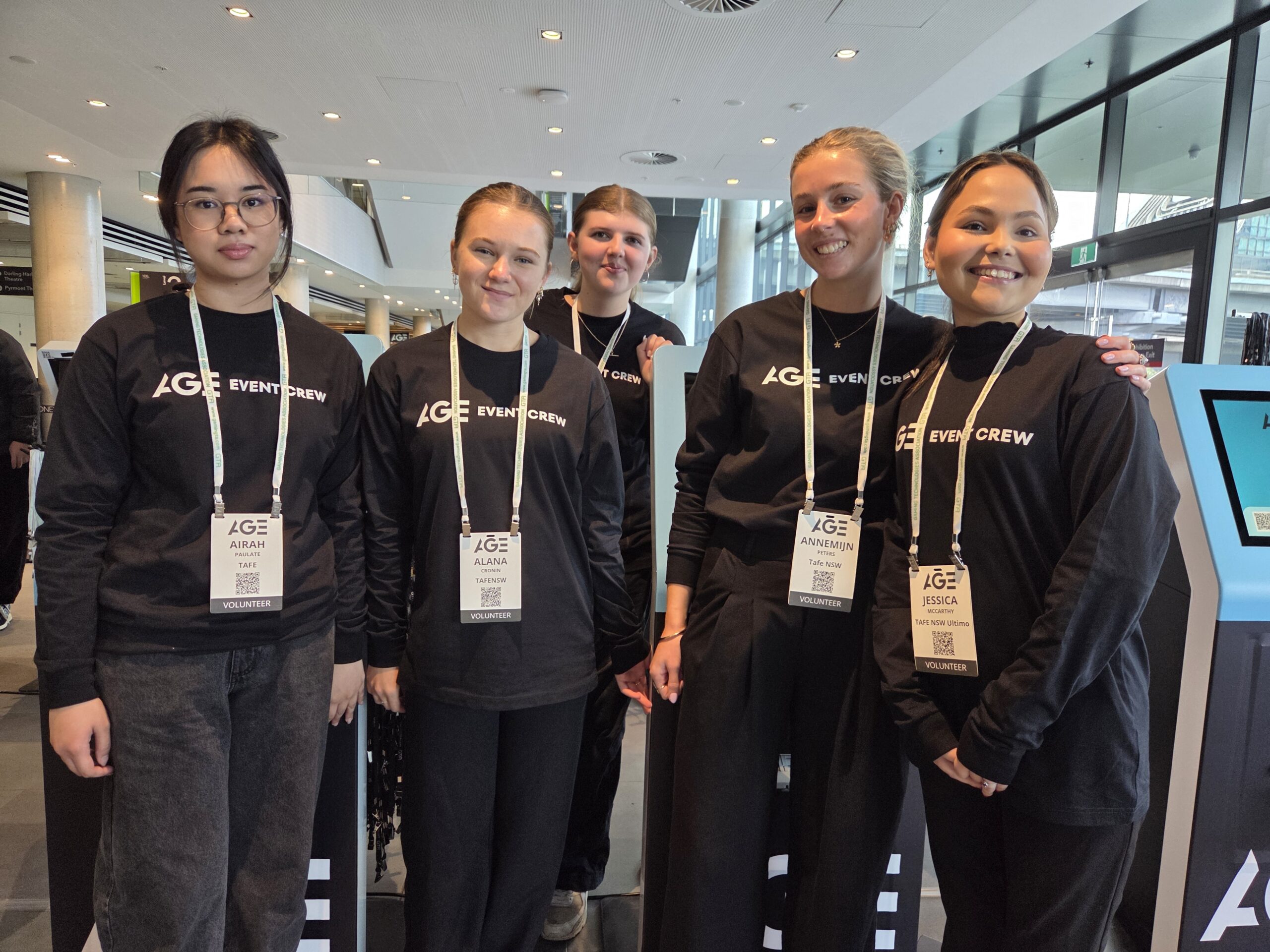Five young women in black AGE EVENT CREW shirts smile together at a gaming 2025 event indoors.