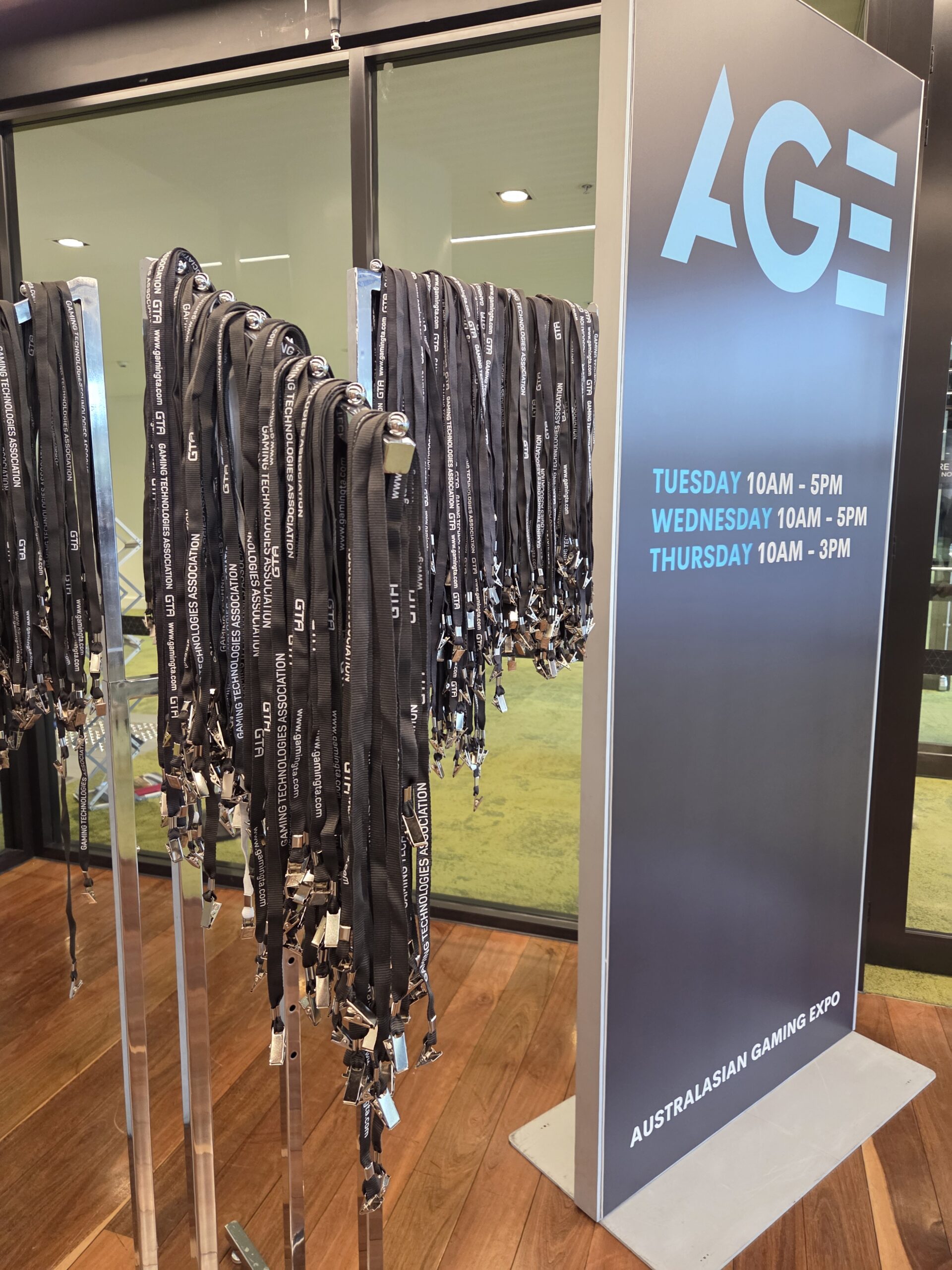 Lanyards hang by an Australasian Gaming Expo sign listing event days, times, and gaming 2025.