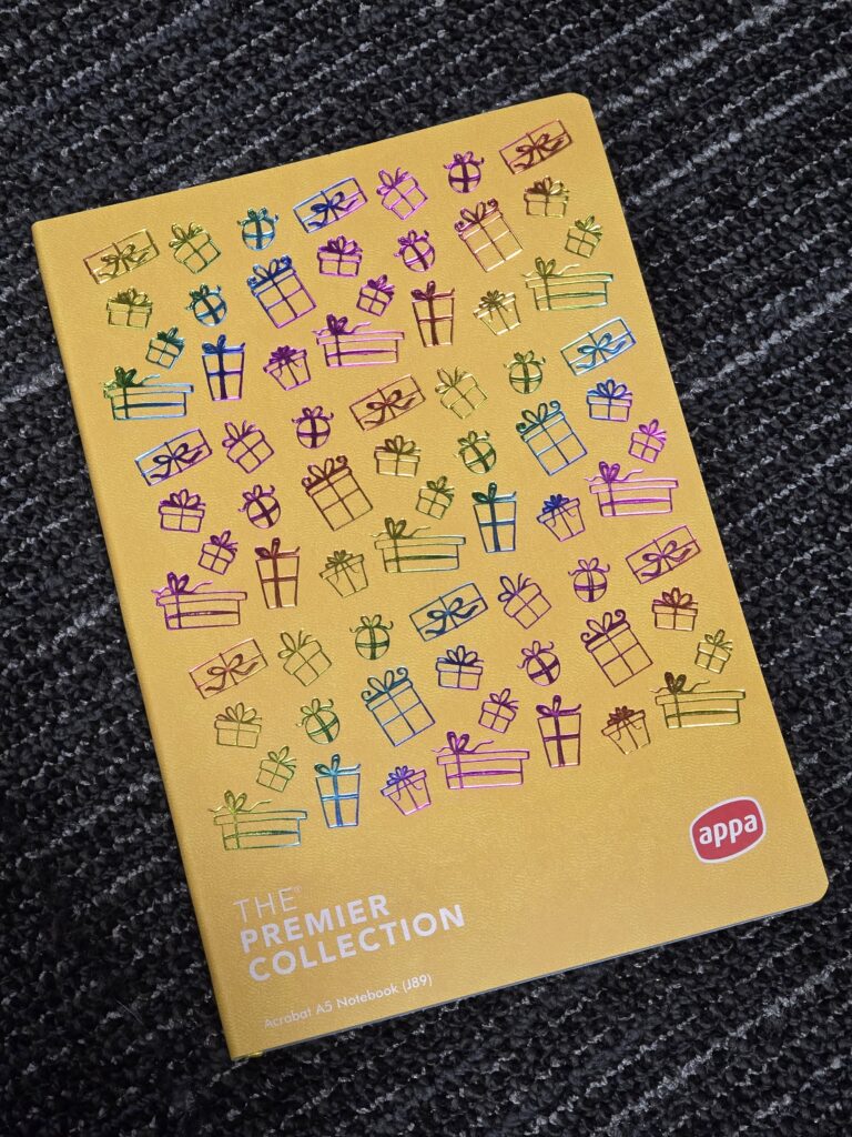 Yellow branded notebook with colorful gift box illustrations on a dark patterned carpet.