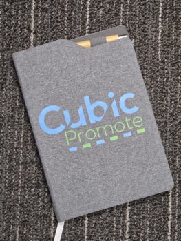 Gray branding notebook with Cubic Promote logo, holding a pen, on a textured carpet.