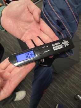 Digital luggage scale reading 10.37 kg held over a striped sleeve and some umbrellas.