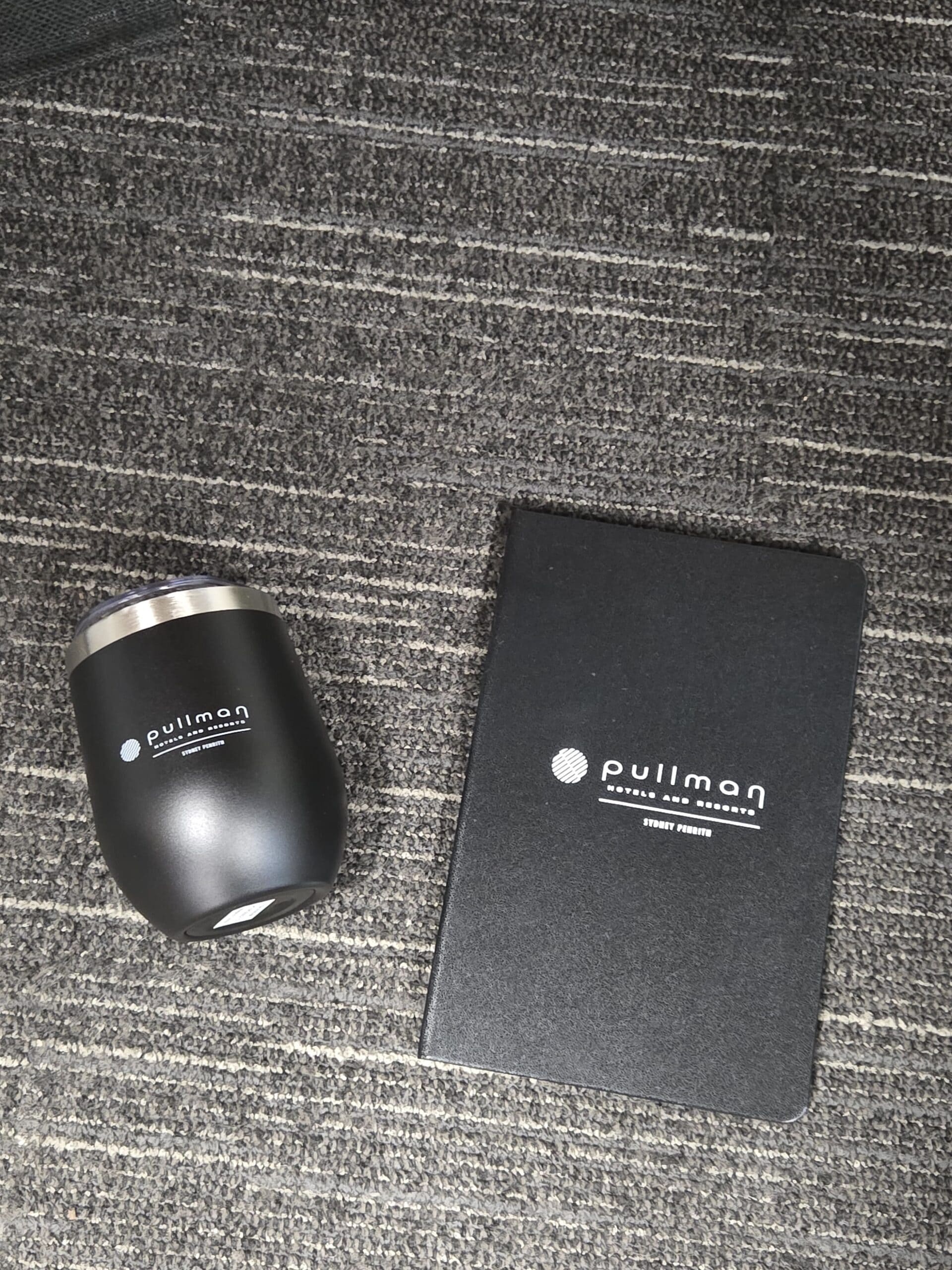 Black tumbler and notebook with Pullman Hotels Sydney logos on a gray carpeted floor.