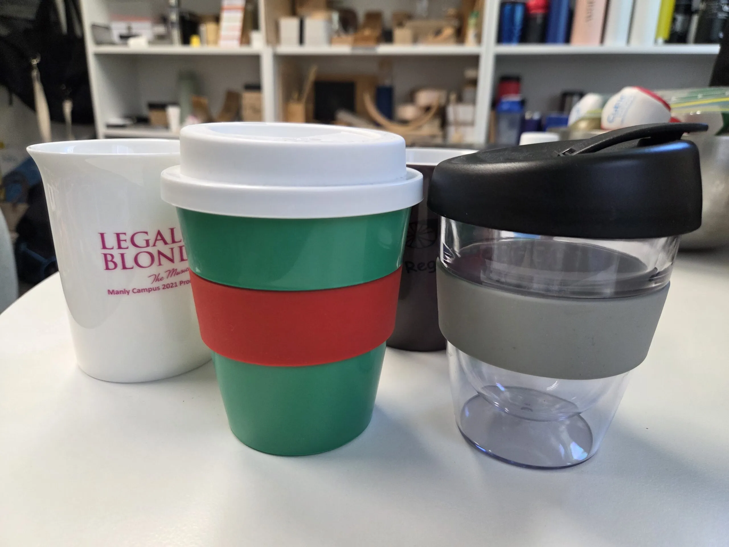 Four reusable promotional mugs in various colors and styles are displayed on a white table.