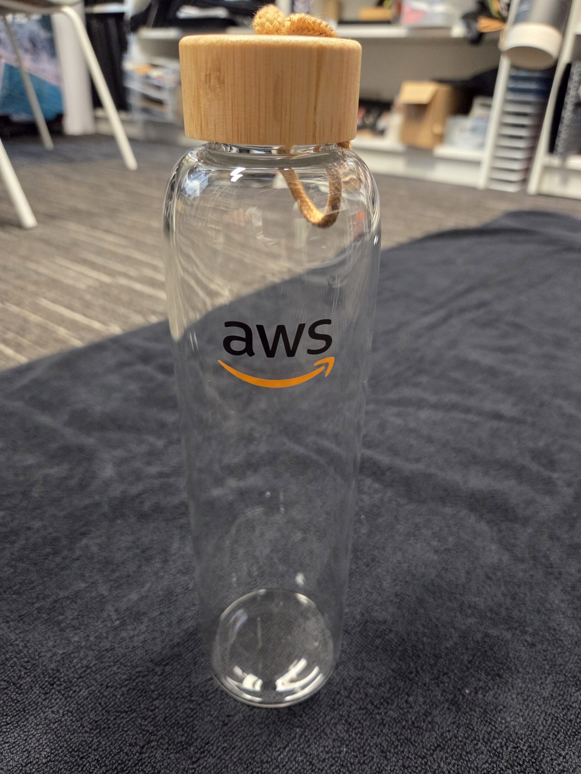 Clear glass which bottle with bamboo lid and aws logo, placed on a dark cloth surface.