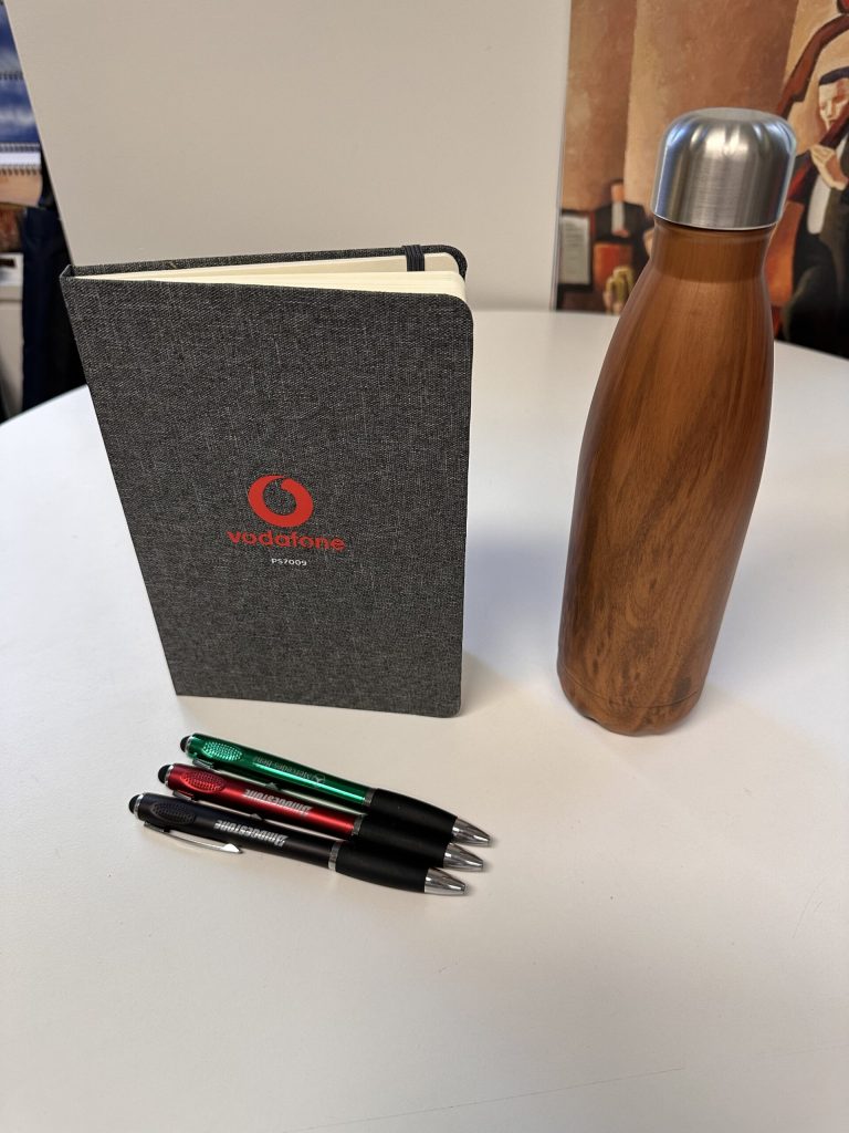 Promotional products: a notebook, wooden water bottle, and four pens on a white table.