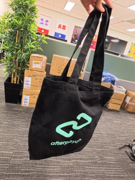 A black Afterpay rebrand tote bag held in an office with boxes and a plant in the background.