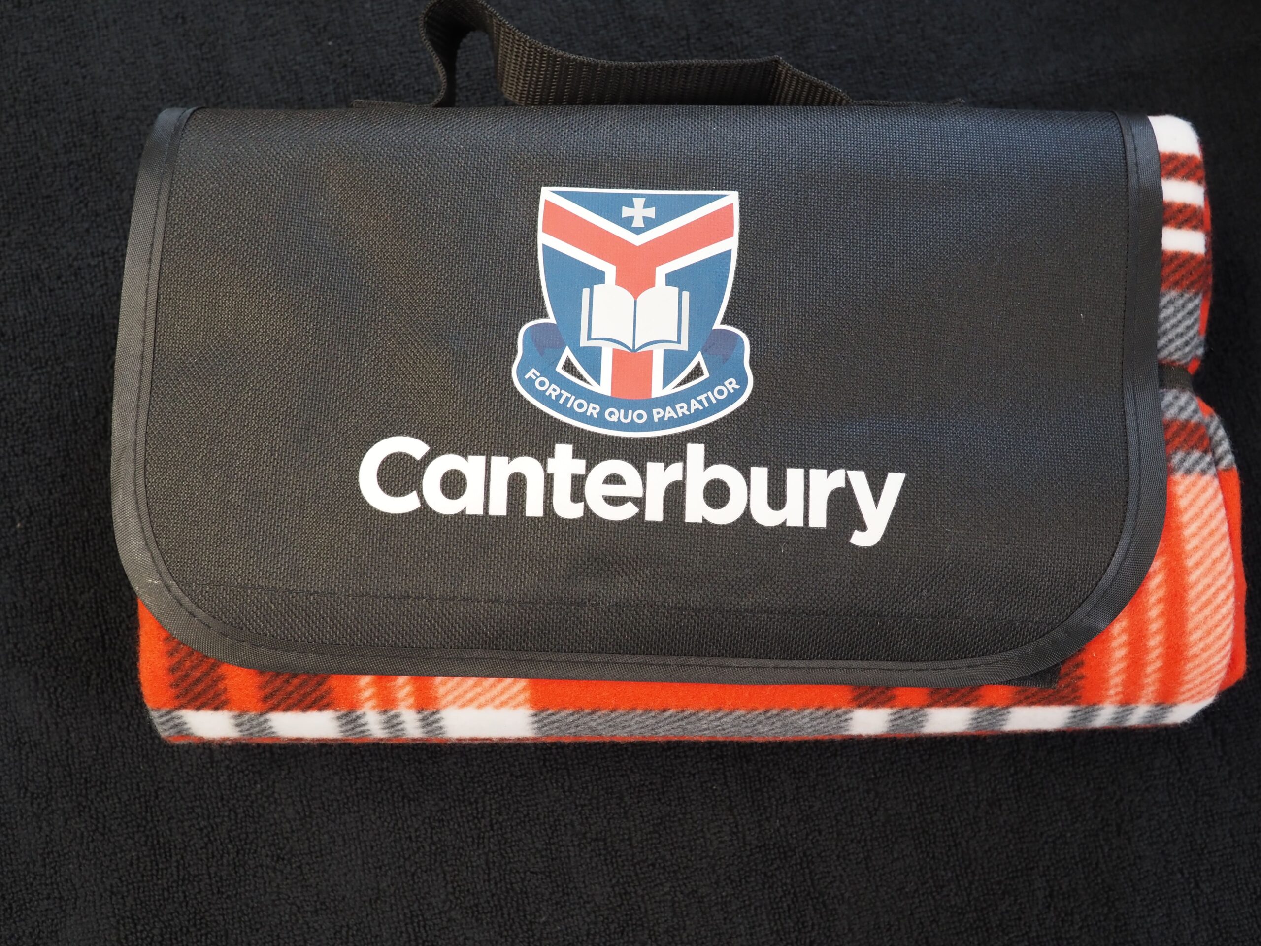 A black Canterbury College blanket bag with a crest and red plaid blanket underneath.