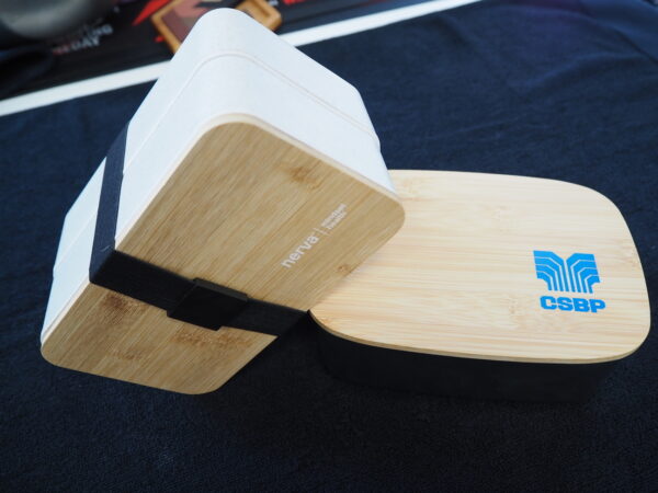 Two rectangular bamboo lunch boxes, ideal as corporate gifts, on a dark fabric surface.
