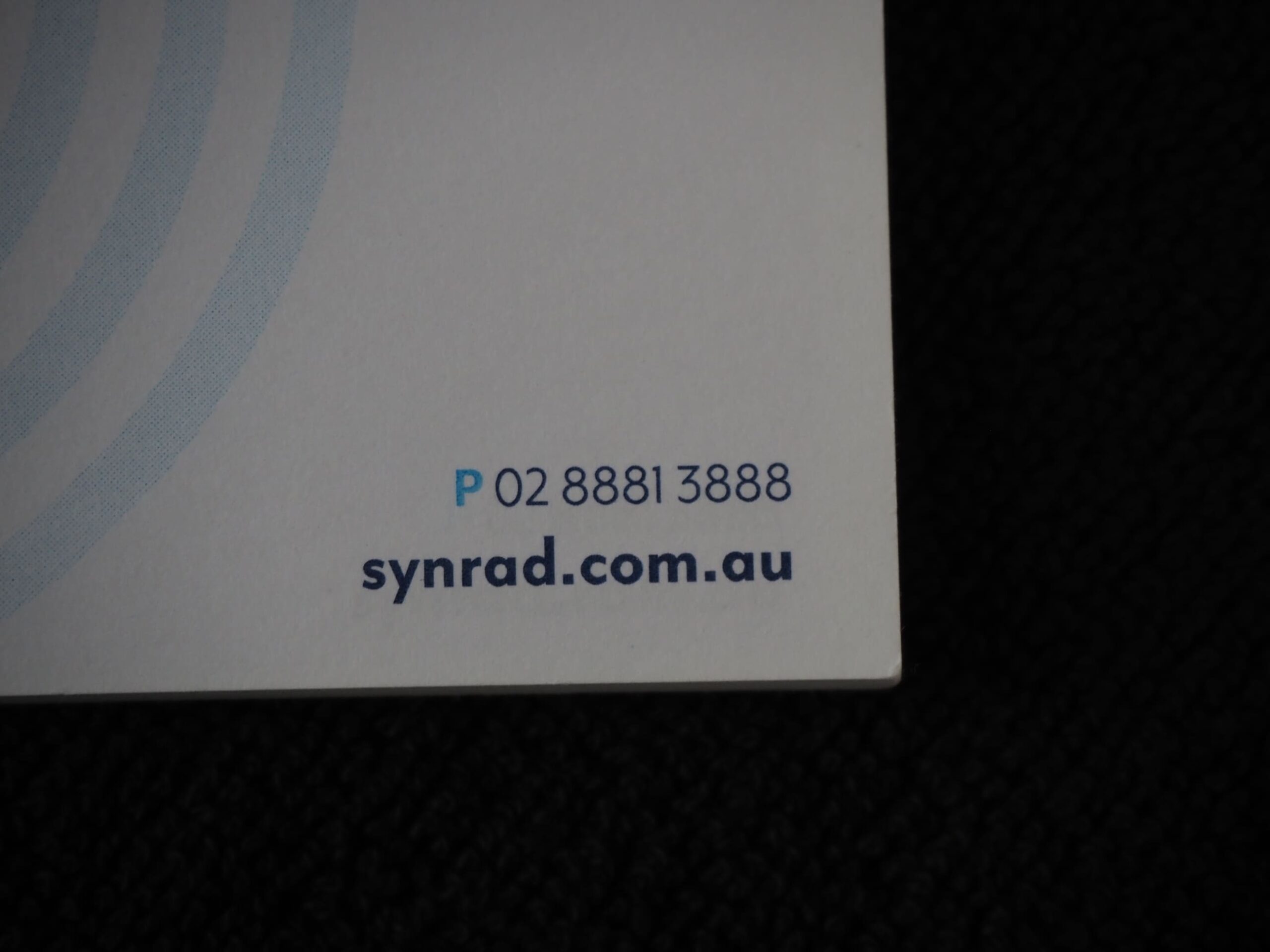 Corner of a card showing Synergy Radiology, 02 8881 3888, and synrad.com.au on a dark surface.