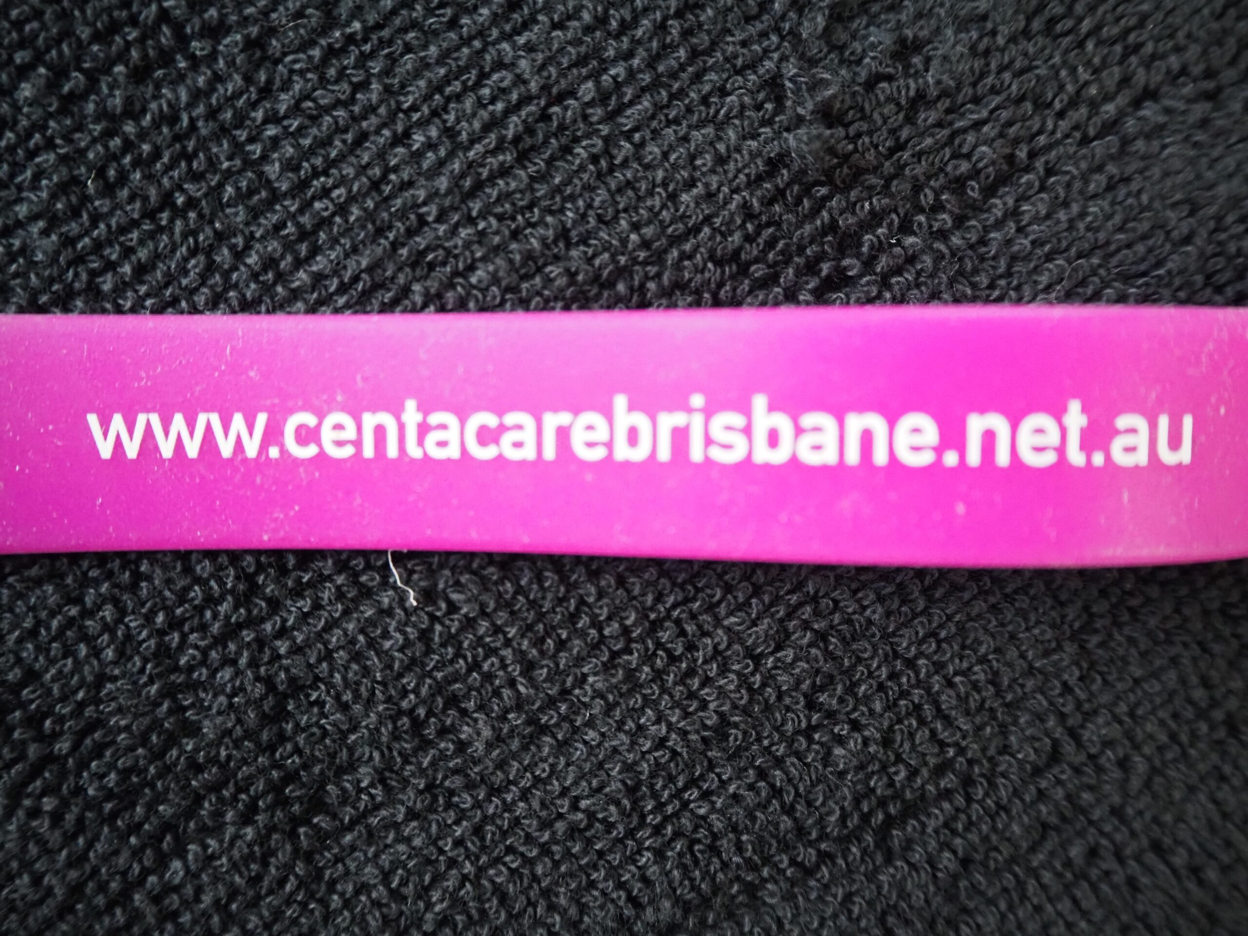 Pink wristband with Centacare website, www.centacarebrisbane.net.au, on black fabric background.