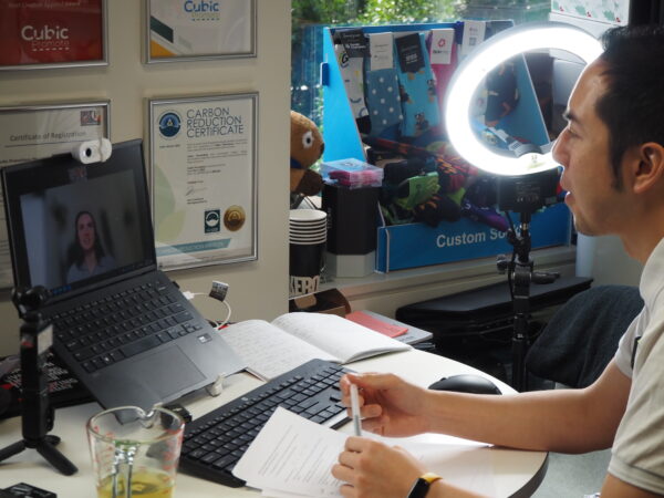 Man in a video call at desk, surrounded by papers, laptop, and branded promotional products.