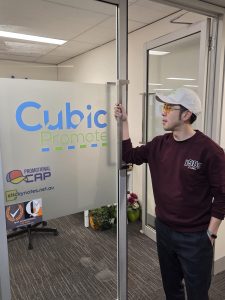 Man in sunglasses and cap stands at Cubic Promote office, specializing in promotional merchandise.