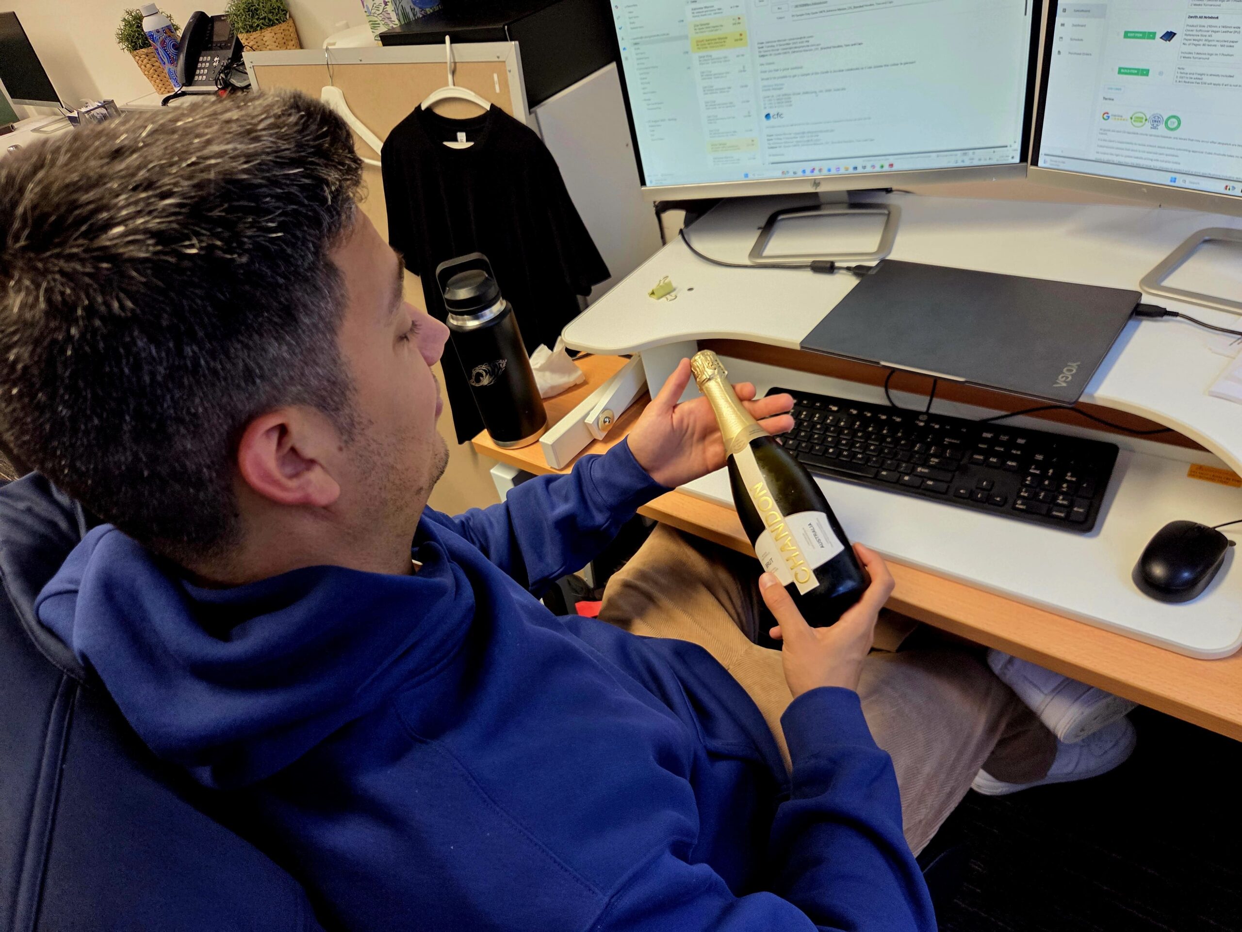 Person in blue hoodie at desk with champagne bottle, working for a promotional products supplier.