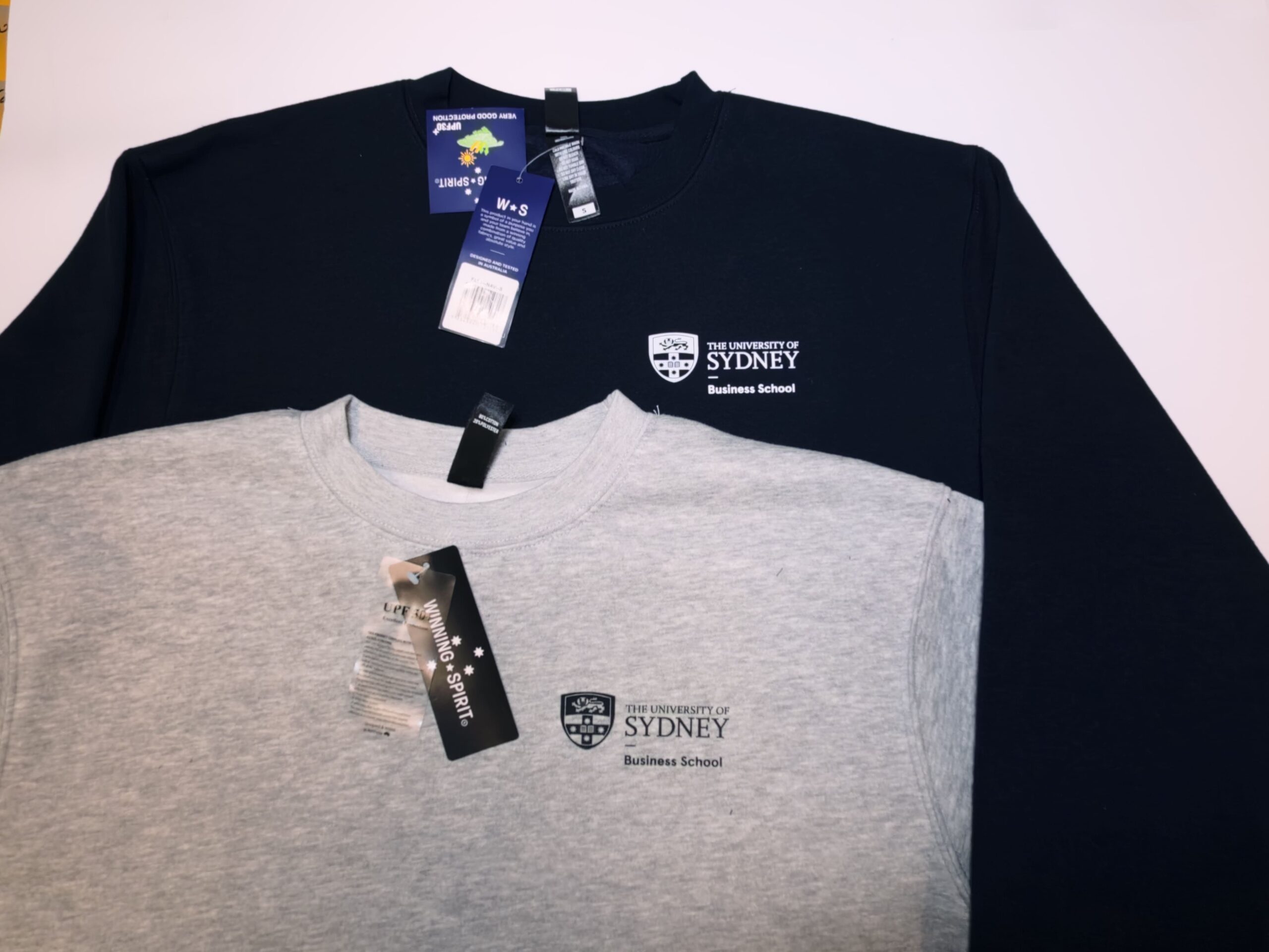 Two University of Sydney Business School sweatshirts, navy and grey, with showroom tags.