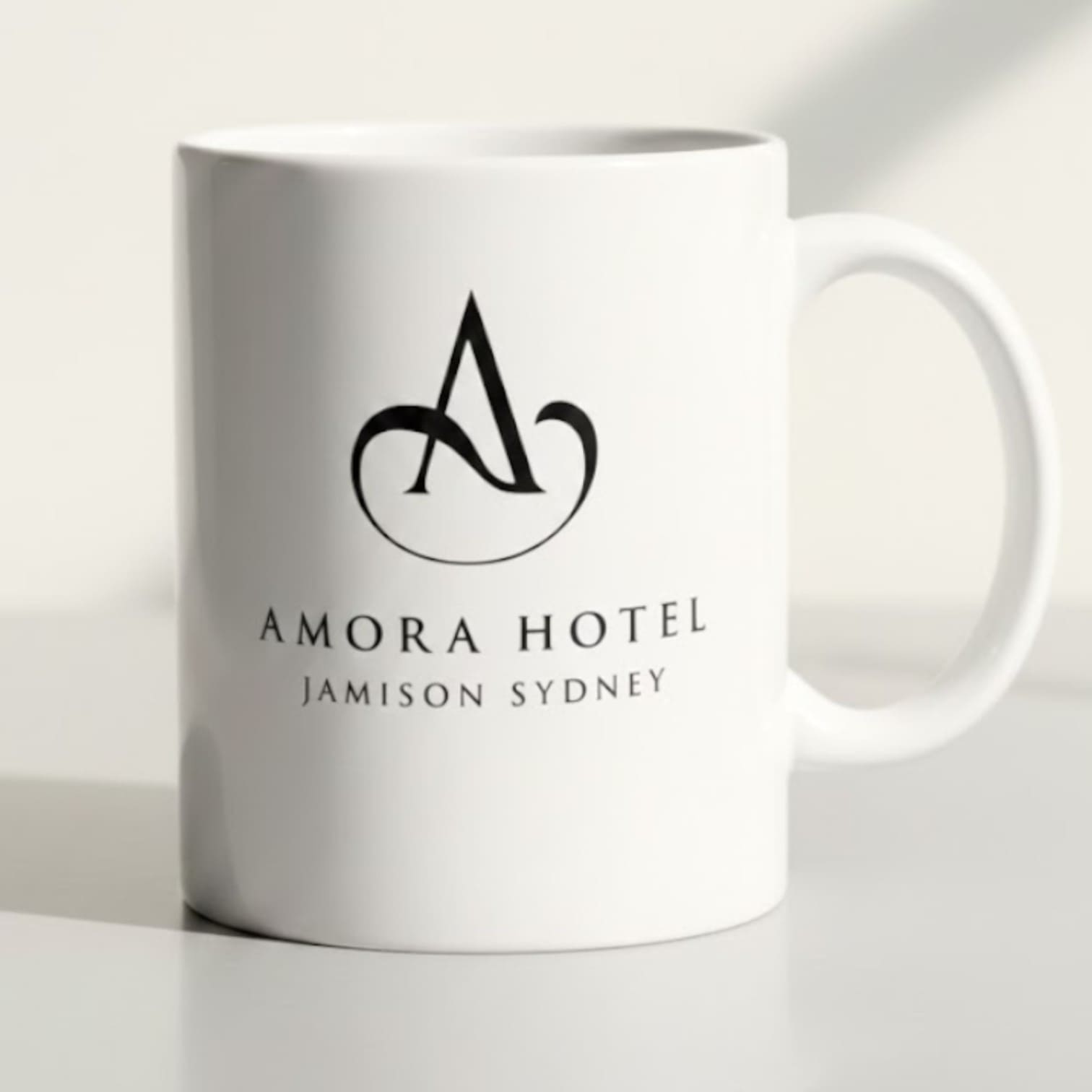 White mug, perfect for any starter kit, with Amora Hotel Jamison Sydney and stylized A logo in black.