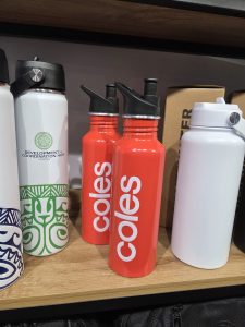 Two red water bottles with Coles branding beside other bottles on a wooden shelf.