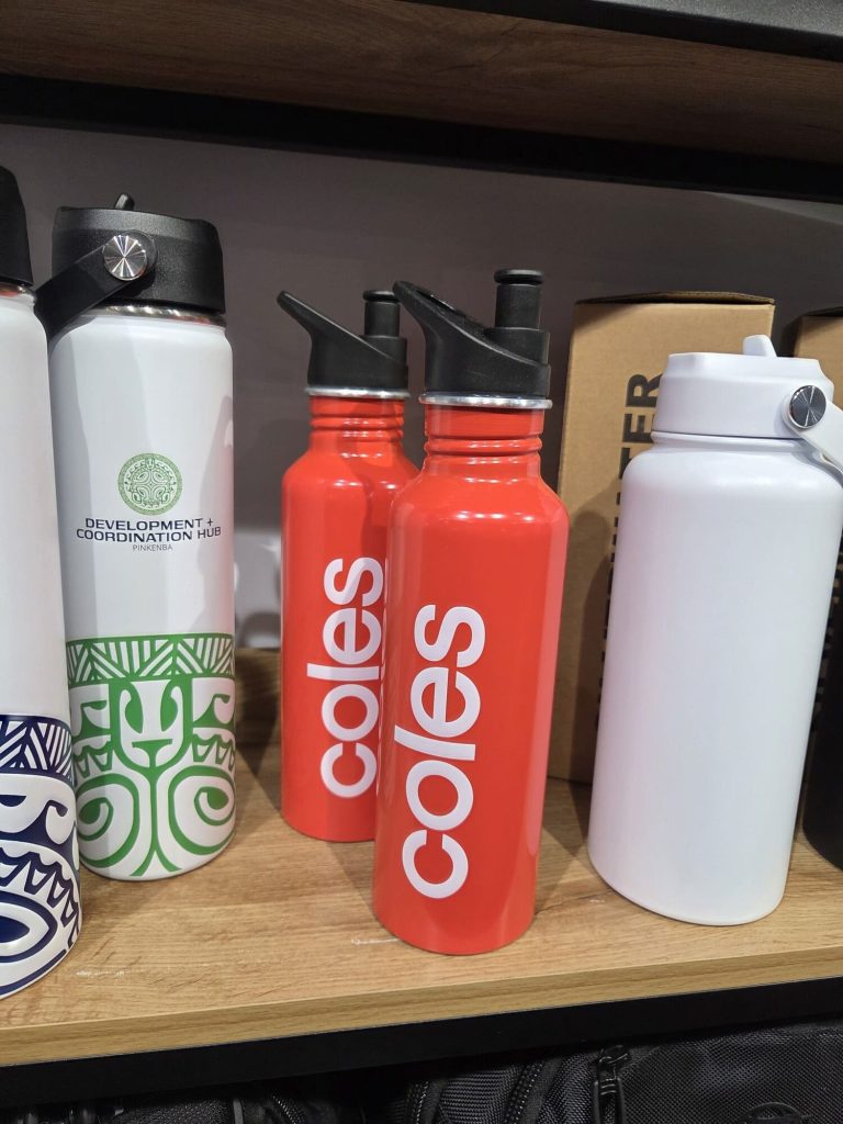 Two red water bottles with Coles branding beside other bottles on a wooden shelf.