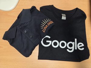 Two black shirts on a desk, each with screen printing: Google text and Luna Park sunburst logo.