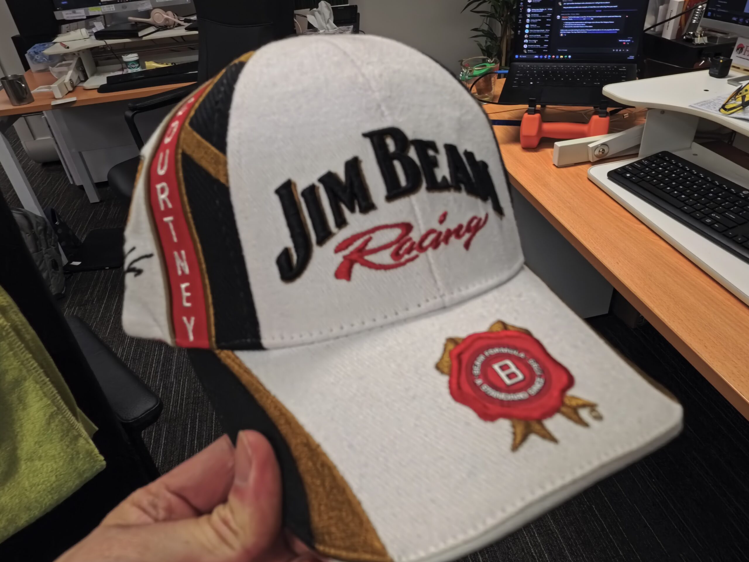 A hand holds a white Jim Beam Racing cap, showcasing expert product sourcing in an office.