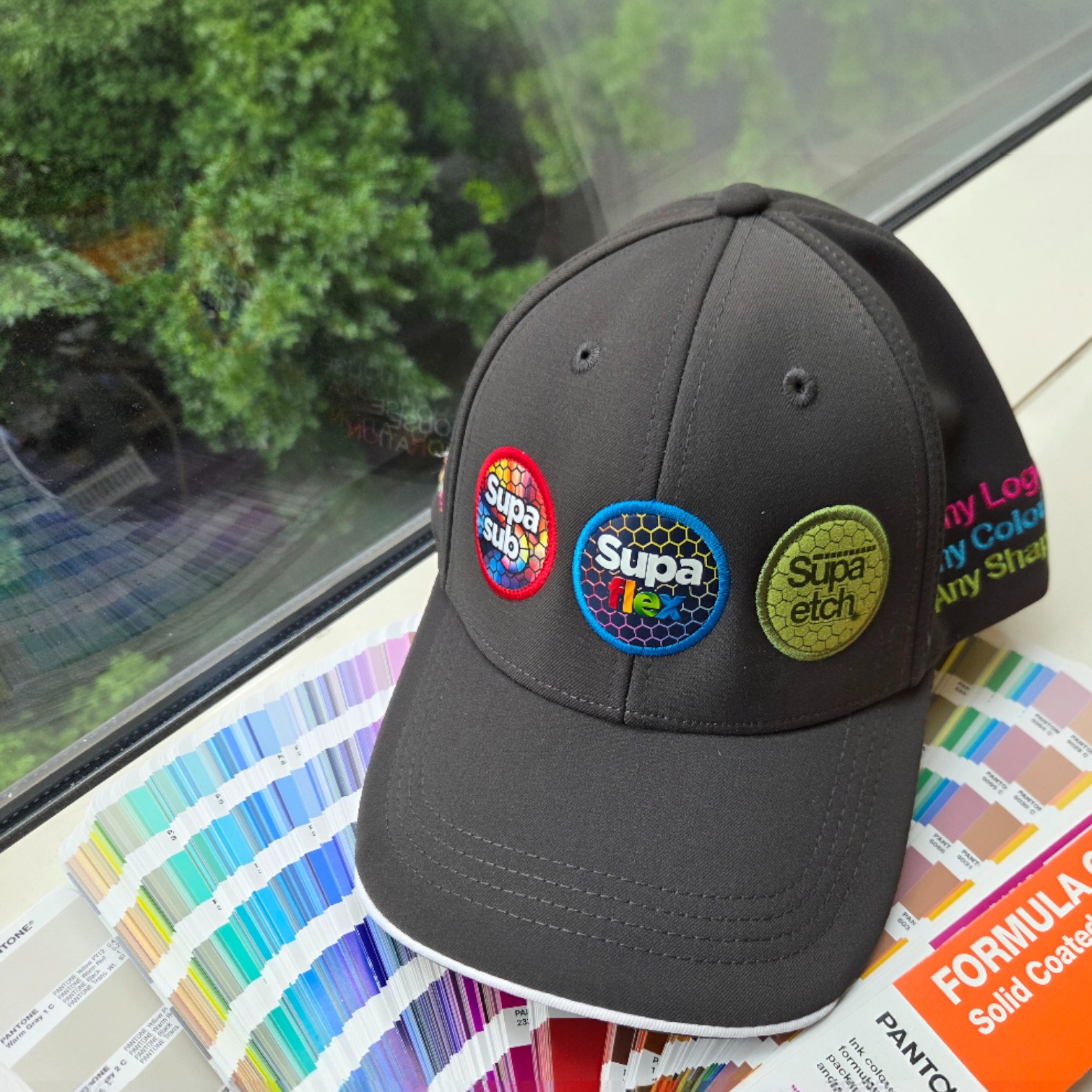 Gray cap with three colorful patches, highlighting print colour accuracy by the window.