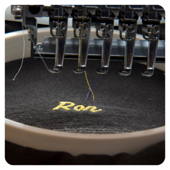 Close-up of an embroidery machine stitching "Ron" on black fabric for durable golf shirts.