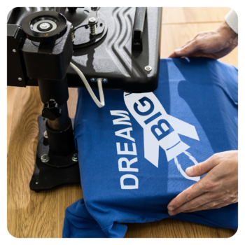 Hands pressing a blue golf shirt with DREAM BIG and a rocket using a heat press machine.