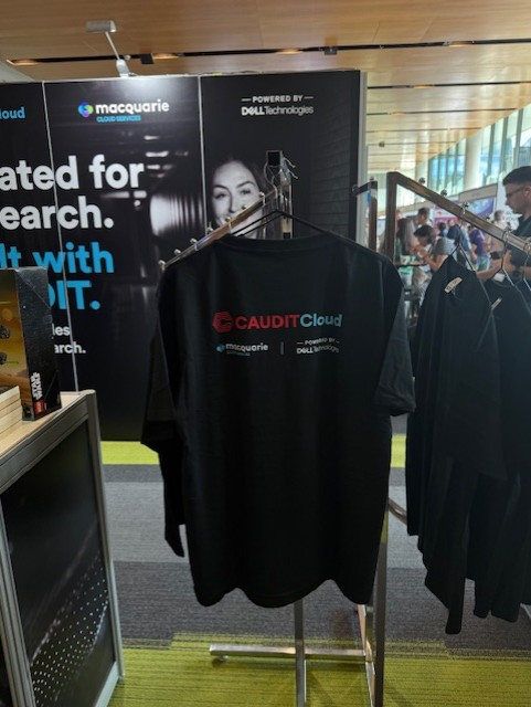 Black CAUDIT Cloud t-shirt on a rack, with Macquarie Cloud Services banners in the background.