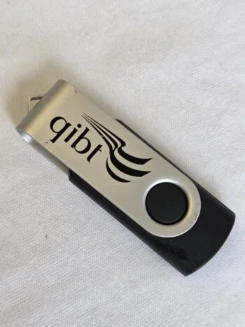 A black and silver Cap USB Drive labeled qibt rests on a white fabric surface.