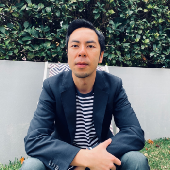 Meet the team: Man in a striped shirt and blazer sitting on grass in front of a green hedge.