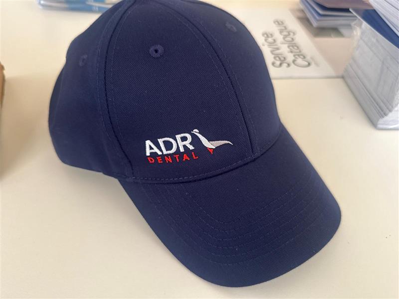 Navy blue cap featuring ADR Dental logo, bird graphic, and a nod to our Client Story.
