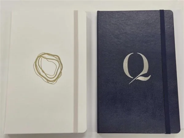 two closed notebooks, perfect as event merchandise, one with a gold swirl and one with a silver Q.